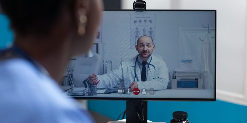 African american asisstant discussing flu symptoms with remote physician doctor during online videocall meeting conference working in hospital office. Telemedicine call on computer screen