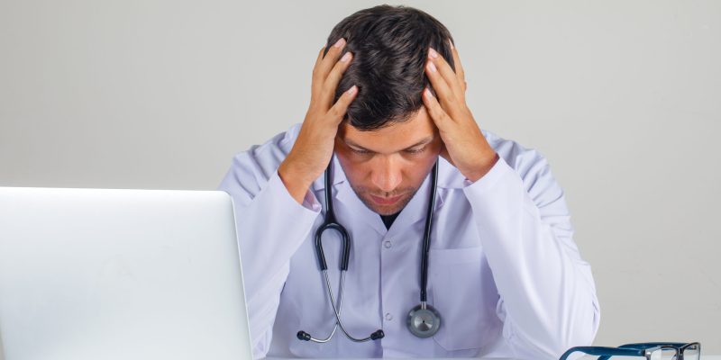 Doctor holding his head in his hands in white coat, stethoscope and looking thoughtful , front view.