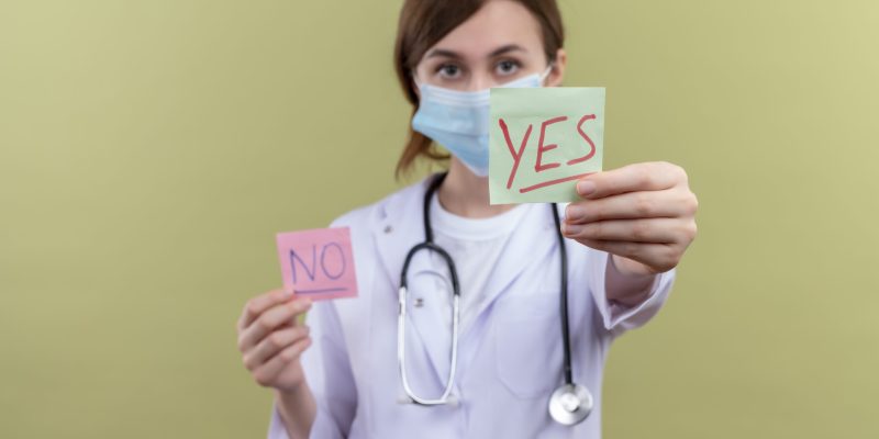 young female doctor wearing medical robe, mask and stethoscope stretching yes note at camera on isolated green background with copy space